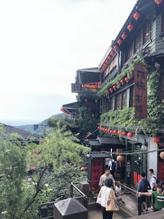 A charming hillside building adorned with red lanterns and green ivy is bustling with visitors. The architecture reveals traditional design elements with multiple stories and balconies. Visitors are seen walking along the pathway that runs alongside this building, surrounded by lush greenery and a distant view of mountains under a slightly overcast sky.