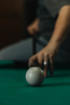 A close-up shot captures a white billiard ball on a green pool table, with a person&rsquo;s hand poised in the background holding a cue stick, indicating a moment of concentration and precision.