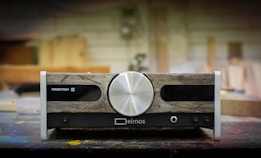 A piece of audio equipment with a vintage aesthetic, featuring a large central silver knob. The front panel combines rustic wooden textures with metallic and black elements, displaying brand names 'Erzetich' and 'Deimos'. It rests on a workshop table, with a blurred background suggesting a woodshop environment.