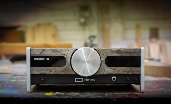 A piece of audio equipment with a vintage aesthetic, featuring a large central silver knob. The front panel combines rustic wooden textures with metallic and black elements, displaying brand names 'Erzetich' and 'Deimos'. It rests on a workshop table, with a blurred background suggesting a woodshop environment.