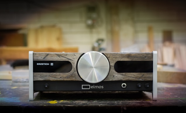 A piece of audio equipment with a vintage aesthetic, featuring a large central silver knob. The front panel combines rustic wooden textures with metallic and black elements, displaying brand names 'Erzetich' and 'Deimos'. It rests on a workshop table, with a blurred background suggesting a woodshop environment.