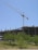 A large construction site with a crane towering over a partially built concrete structure. Several floors are visible, with metal reinforcements sticking out. The foreground includes green trees and grass under a clear blue sky.