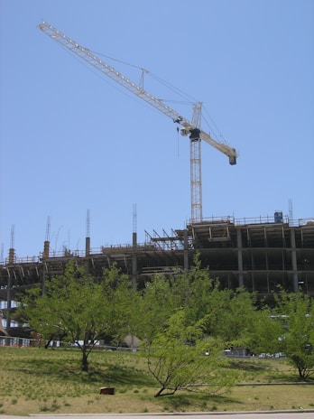 A large construction site with a crane towering over a partially built concrete structure. Several floors are visible, with metal reinforcements sticking out. The foreground includes green trees and grass under a clear blue sky.