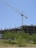 A large construction site with a crane towering over a partially built concrete structure. Several floors are visible, with metal reinforcements sticking out. The foreground includes green trees and grass under a clear blue sky.