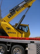 A large yellow crane with the lettering 'Grove' is positioned on a vehicle platform. A red caution tape with the words 'Danger Do Not Enter' is surrounding the area. The sky is clear in the background.