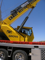A large yellow crane with the lettering 'Grove' is positioned on a vehicle platform. A red caution tape with the words 'Danger Do Not Enter' is surrounding the area. The sky is clear in the background.