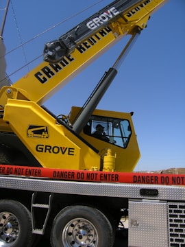 A large yellow crane with the lettering 'Grove' is positioned on a vehicle platform. A red caution tape with the words 'Danger Do Not Enter' is surrounding the area. The sky is clear in the background.
