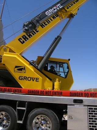 A large yellow crane with the lettering 'Grove' is positioned on a vehicle platform. A red caution tape with the words 'Danger Do Not Enter' is surrounding the area. The sky is clear in the background.