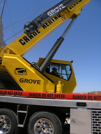A large yellow crane with the lettering 'Grove' is positioned on a vehicle platform. A red caution tape with the words 'Danger Do Not Enter' is surrounding the area. The sky is clear in the background.
