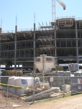 A construction site with an unfinished multi-story building. Metal scaffolding is visible around the structure, and a tall crane stands nearby. The foreground shows construction materials such as pallets, bags, and machinery behind a metal fence with no trespassing signs.