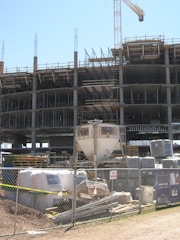 A construction site with an unfinished multi-story building. Metal scaffolding is visible around the structure, and a tall crane stands nearby. The foreground shows construction materials such as pallets, bags, and machinery behind a metal fence with no trespassing signs.