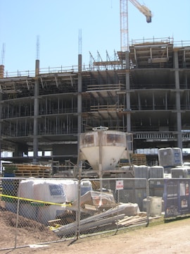 A construction site with an unfinished multi-story building. Metal scaffolding is visible around the structure, and a tall crane stands nearby. The foreground shows construction materials such as pallets, bags, and machinery behind a metal fence with no trespassing signs.
