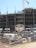 A construction site with an unfinished multi-story building. Metal scaffolding is visible around the structure, and a tall crane stands nearby. The foreground shows construction materials such as pallets, bags, and machinery behind a metal fence with no trespassing signs.