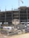A construction site with an unfinished multi-story building. Metal scaffolding is visible around the structure, and a tall crane stands nearby. The foreground shows construction materials such as pallets, bags, and machinery behind a metal fence with no trespassing signs.