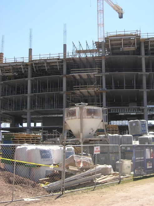 A construction site with an unfinished multi-story building. Metal scaffolding is visible around the structure, and a tall crane stands nearby. The foreground shows construction materials such as pallets, bags, and machinery behind a metal fence with no trespassing signs.