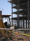 A construction site with a partially built multi-story building featuring scaffolding and metal beams. In the foreground, there is a yellow construction vehicle behind a chain-link fence. A sign for a fencing company is attached to the fence.