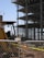 A construction site with a partially built multi-story building featuring scaffolding and metal beams. In the foreground, there is a yellow construction vehicle behind a chain-link fence. A sign for a fencing company is attached to the fence.