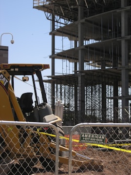 A construction site with a partially built multi-story building featuring scaffolding and metal beams. In the foreground, there is a yellow construction vehicle behind a chain-link fence. A sign for a fencing company is attached to the fence.