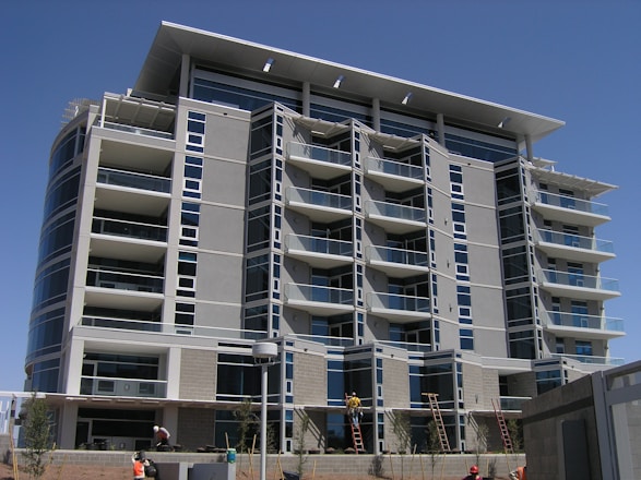 A modern multi-story building under construction with several floors featuring large windows and balconies. Workers and ladders are visible at the ground level, indicating ongoing construction or maintenance work. The architecture includes clean lines and a mix of concrete and glass materials.