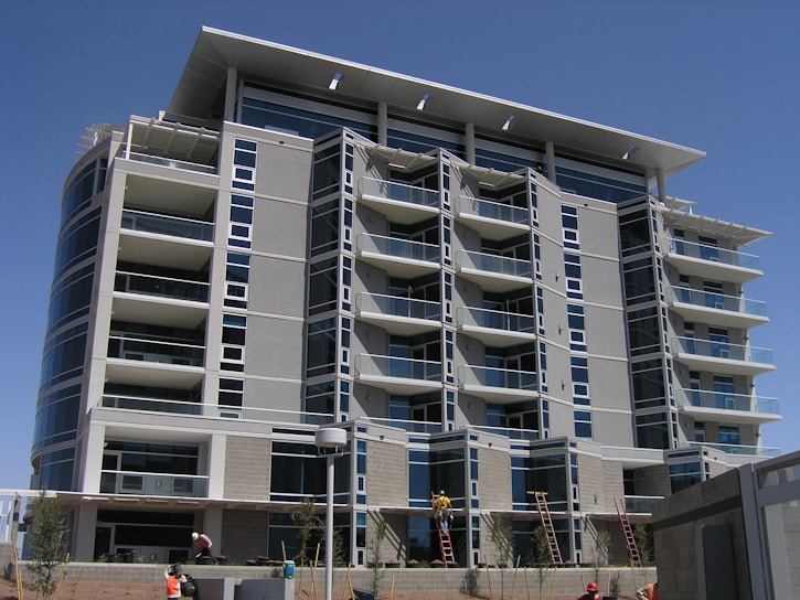 A modern multi-story building under construction with several floors featuring large windows and balconies. Workers and ladders are visible at the ground level, indicating ongoing construction or maintenance work. The architecture includes clean lines and a mix of concrete and glass materials.