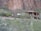 A large adobe-style house is nestled in a desert landscape surrounded by cacti and brush. The exterior has earthy tones with rounded corners and wooden accents. There is a rocky, mountainous backdrop and greenery in the foreground.