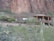 A large adobe-style house is nestled in a desert landscape surrounded by cacti and brush. The exterior has earthy tones with rounded corners and wooden accents. There is a rocky, mountainous backdrop and greenery in the foreground.