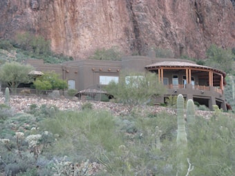 A large adobe-style house is nestled in a desert landscape surrounded by cacti and brush. The exterior has earthy tones with rounded corners and wooden accents. There is a rocky, mountainous backdrop and greenery in the foreground.