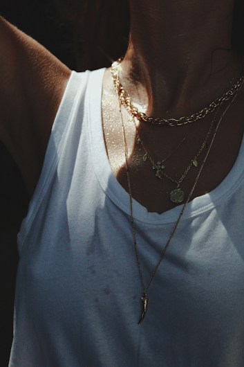 A close-up view of a person wearing multiple gold necklaces over a white tank top. The lighting is soft, with a slight shimmer on the skin, creating a sense of warmth.
