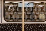 The interior of a subway train window with patterned illustrations of a landmark, possibly the Brandenburg Gate. The window display includes a small advertisement featuring a creative space explorer and a sign with a video camera icon. The train seat shows a dark, abstract pattern with small colored details.