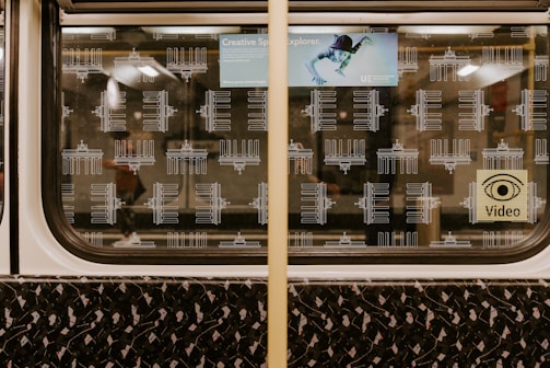 The interior of a subway train window with patterned illustrations of a landmark, possibly the Brandenburg Gate. The window display includes a small advertisement featuring a creative space explorer and a sign with a video camera icon. The train seat shows a dark, abstract pattern with small colored details.