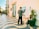 A person is painting on an easel outdoors on a tiled, wave-patterned sidewalk. Nearby, there are potted plants, chairs, and an iron gate attached to a building. The painting depicts a waterfront scene.