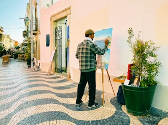 A person is painting on an easel outdoors on a tiled, wave-patterned sidewalk. Nearby, there are potted plants, chairs, and an iron gate attached to a building. The painting depicts a waterfront scene.