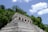 A stone pyramid structure with multiple levels, partially concealed by dense green foliage and set against a clear blue sky with fluffy clouds. The pyramid, likely an ancient ruin, displays intricate stonework and steps leading to a small structure at the top.