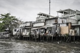 A row of makeshift houses built on stilts lines the edge of a murky body of water. The structures appear to be made of corrugated metal, bricks, and wood, with some sections covered by tarpaulins. Various items, such as clothing, hang outside to dry. Behind these homes, more modern white buildings with balconies can be seen. Vegetation, including a large leafy tree, is present in the background under a cloudy sky.