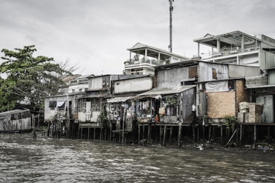 A row of makeshift houses built on stilts lines the edge of a murky body of water. The structures appear to be made of corrugated metal, bricks, and wood, with some sections covered by tarpaulins. Various items, such as clothing, hang outside to dry. Behind these homes, more modern white buildings with balconies can be seen. Vegetation, including a large leafy tree, is present in the background under a cloudy sky.