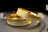 Two gold bracelets with intricate textures and patterns are lying on a dark surface. The bracelets have a detailed, textured design along their bands, reflecting light and displaying a glossy finish.