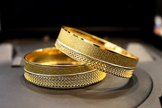Two gold bracelets with intricate textures and patterns are lying on a dark surface. The bracelets have a detailed, textured design along their bands, reflecting light and displaying a glossy finish.