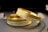Two gold bracelets with intricate textures and patterns are lying on a dark surface. The bracelets have a detailed, textured design along their bands, reflecting light and displaying a glossy finish.