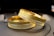 Two gold bracelets with intricate textures and patterns are lying on a dark surface. The bracelets have a detailed, textured design along their bands, reflecting light and displaying a glossy finish.