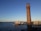 A tall brick chimney stands in the foreground against a clear blue sky. In the distance across a body of water, there is a distinct white architectural structure resembling sails, and a shoreline with buildings.