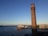 A tall brick chimney stands in the foreground against a clear blue sky. In the distance across a body of water, there is a distinct white architectural structure resembling sails, and a shoreline with buildings.