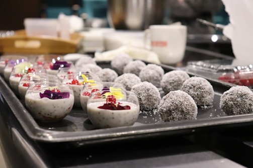A tray is filled with round, coconut-covered desserts and small glass cups containing creamy chia pudding topped with vibrant, colorful flowers and red berries. The setting appears to be a kitchen or a food preparation area with a mug and some kitchen items in the background.