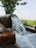 A large pipe pours a steady stream of clear water into a pool, surrounded by stone bricks. The scene is set in a lush, green area with visible grass and a tree providing some shade. There are open fields with a view extending to the horizon under a clear sky.