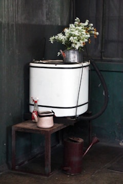 A rustic corner features an old washing machine with a metal watering can holding white and orange flowers placed on top. A small, weathered stool supports another kettle and a spray bottle nearby, suggesting a vintage, utilitarian setting.