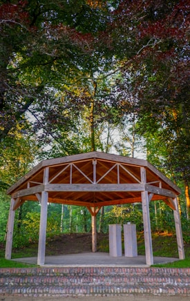 A wooden gazebo is situated in a forested area, surrounded by lush greenery and tall trees. The structure features an open design with visible beams and a tiled roof. In front of the gazebo are two rectangular pedestals.