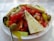 A fresh and vibrant Greek salad topped with large slices of feta cheese, sliced tomatoes, cucumbers, olives, and pepperoncini peppers. The salad is sprinkled with herbs and served on a white plate with a spoon on the side.