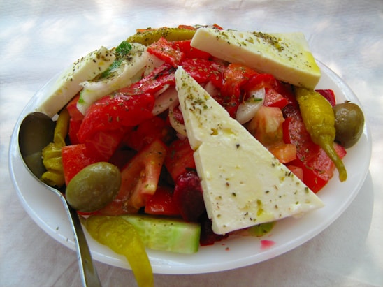 A fresh and vibrant Greek salad topped with large slices of feta cheese, sliced tomatoes, cucumbers, olives, and pepperoncini peppers. The salad is sprinkled with herbs and served on a white plate with a spoon on the side.