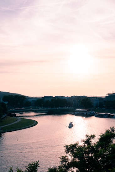 A serene river scene at sunset, with a small boat cruising through calm waters. The sky is filled with soft hues of orange and pink, reflecting onto the water's surface. Silhouettes of buildings and trees line the riverbank.