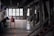 A dimly lit industrial interior with large windows on one side and various metal structures and equipment placed throughout the space. The room has a utilitarian design, featuring exposed beams and a stark ambiance. There is a long red metal conveyor table centered in the image and multiple angled chutes lined along one wall.
