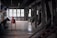 A dimly lit industrial interior with large windows on one side and various metal structures and equipment placed throughout the space. The room has a utilitarian design, featuring exposed beams and a stark ambiance. There is a long red metal conveyor table centered in the image and multiple angled chutes lined along one wall.